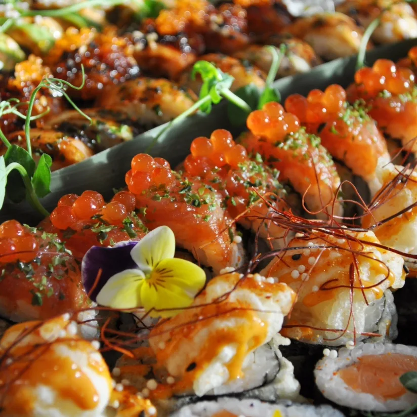 An omakase platter close up: salmon nigiri piled with ikura and herbs, tempura rolls dressed in spicy mayo and chilli threads, torched rolls behind, a pansy on top